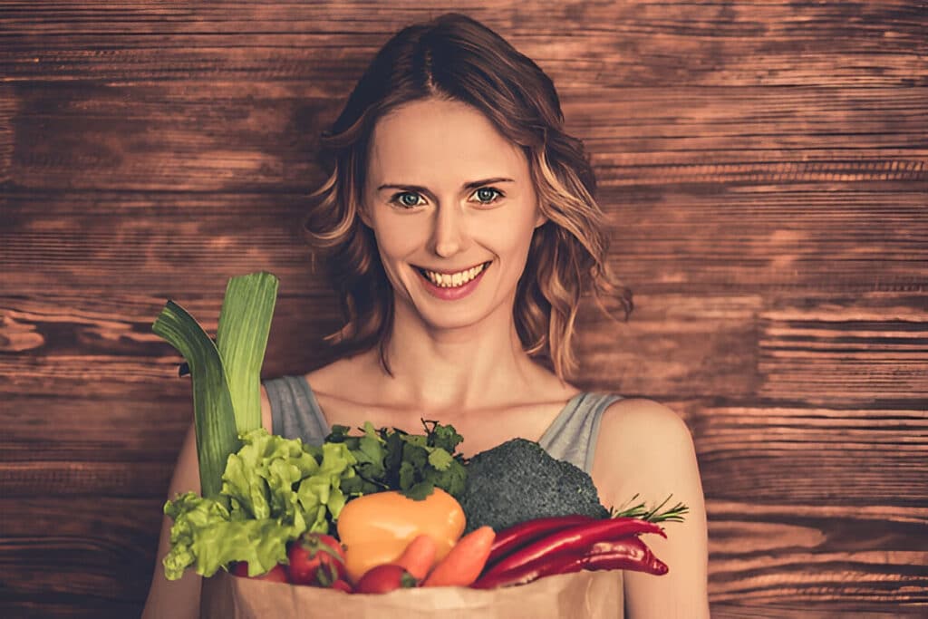 About Me 4 Ivy smiling and holding a paper grocery bag filled with fresh vegetables like leeks, lettuce, bell peppers, and broccoli, showing her love for healthy cooking