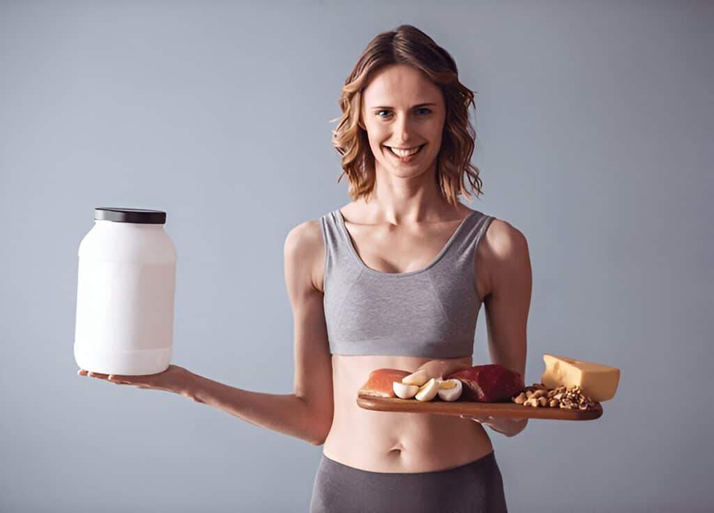 About Me 2 Ivy smiling in fitness clothes, holding a large protein powder container in one hand and a tray of high-protein foods in the other, showing her commitment to a balanced, protein-rich diet
